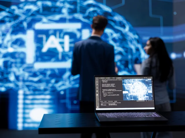 Laptop displaying AI cognitive computing technology, with IT staff members working in the blurry background, highlighting the use of advanced AI tools for cybersecurity