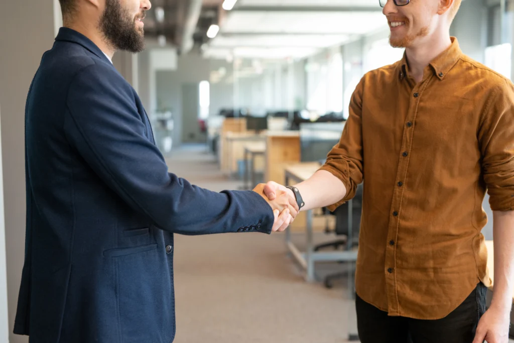 Two young colleagues in shaking hands in an open-space office, symbolizing collaboration and trust in the context of cybersecurity and AI-driven solutions.