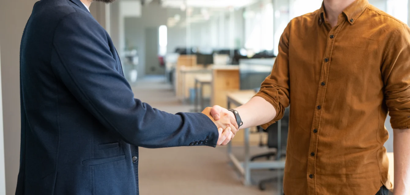 Two young colleagues in shaking hands in an open-space office, symbolizing collaboration and trust in the context of cybersecurity and AI-driven solutions.