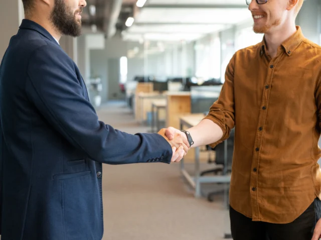 Two young colleagues in shaking hands in an open-space office, symbolizing collaboration and trust in the context of cybersecurity and AI-driven solutions.