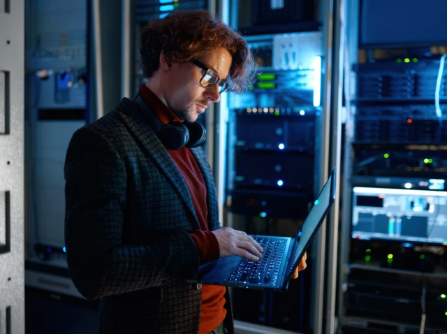IT technician examining server diagnostics on a laptop in a dark server room, highlighting the importance of addressing cybersecurity gaps often overlooked by SMEs